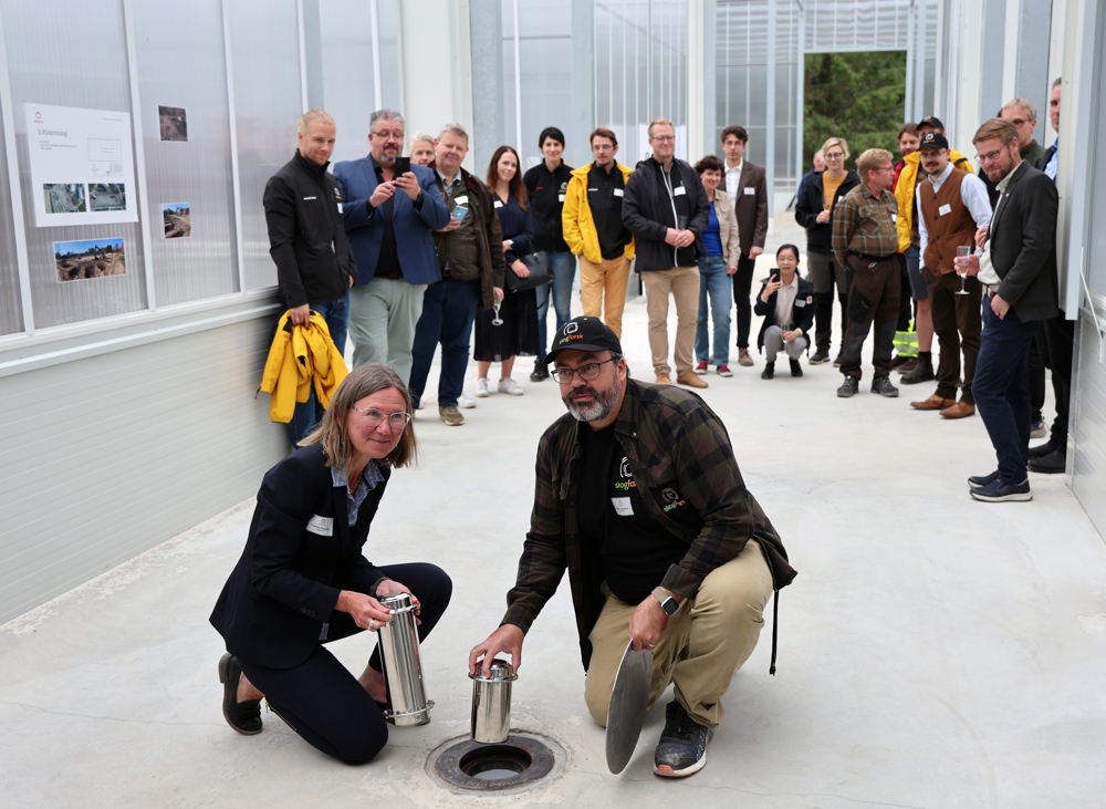 Two persons putting down steel can in floor in greenhouse, in front of group of people. Photo.